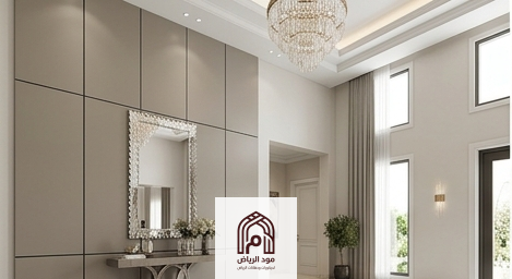 مدخل فيلا فاخر ذو سقف عالي في مدينة الرياض يتميز بجدار كامل من ألواح بديل الشيبورد الناعمة بلون بيج رمادي (جريج) مع فواصل ظل سوداء هندسية ناعمة وإضاءة ثريا كريستال.معلم بديل الشيبورد بالرياض.